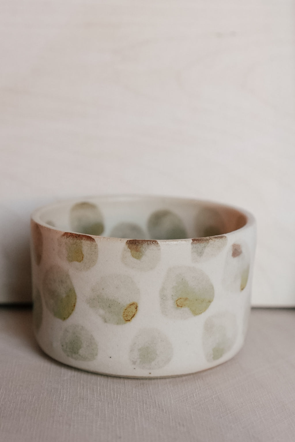 A handmade ceramic cup with a white base and irregular, hand-painted green and brown polka dots, set against a neutral light gray background.