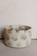 A handmade ceramic cup with a white base and irregular, hand-painted green and brown polka dots, set against a neutral light gray background.