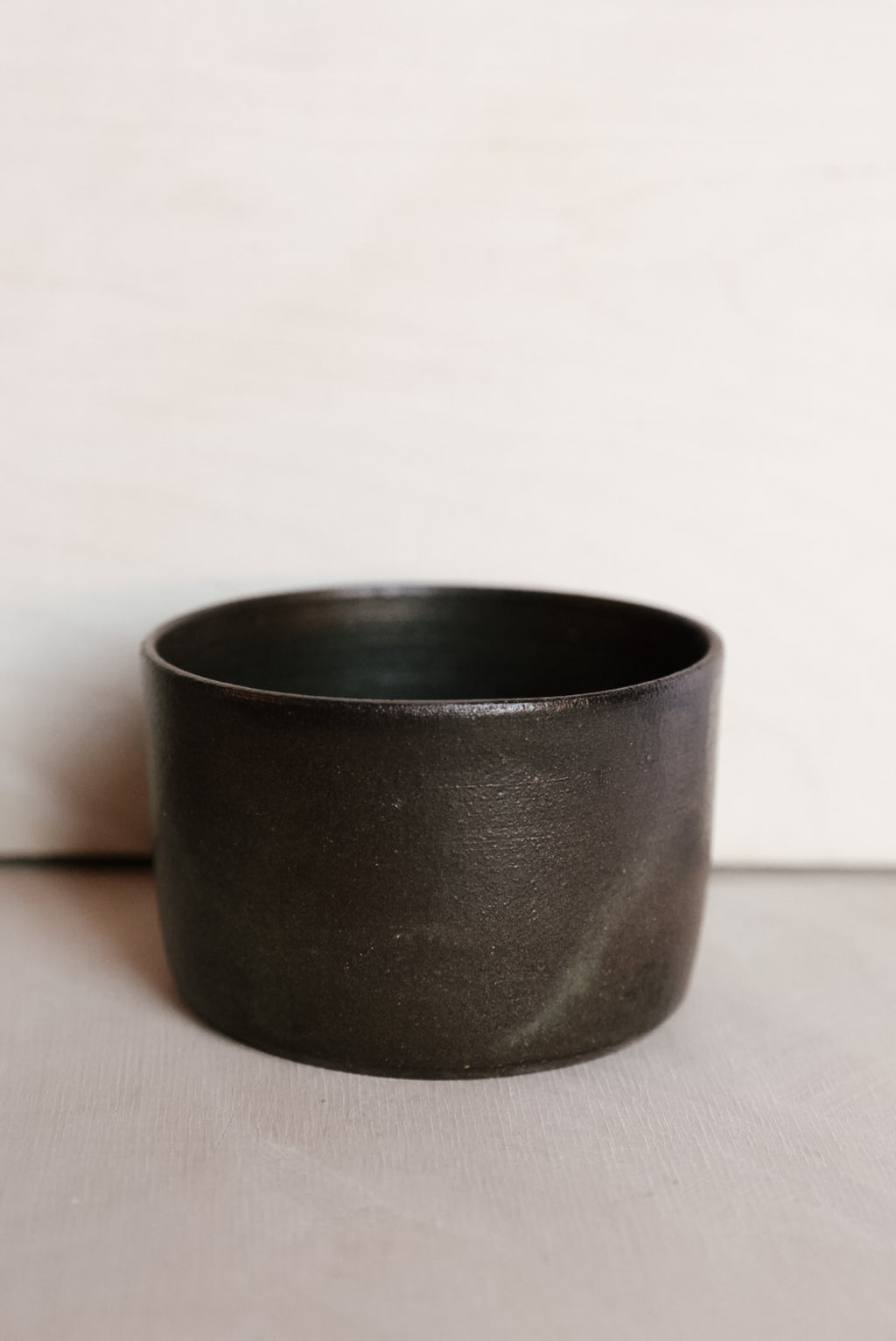A dark charcoal gray - brown ceramic cup with a matte, textured finish, set against a neutral light gray background.