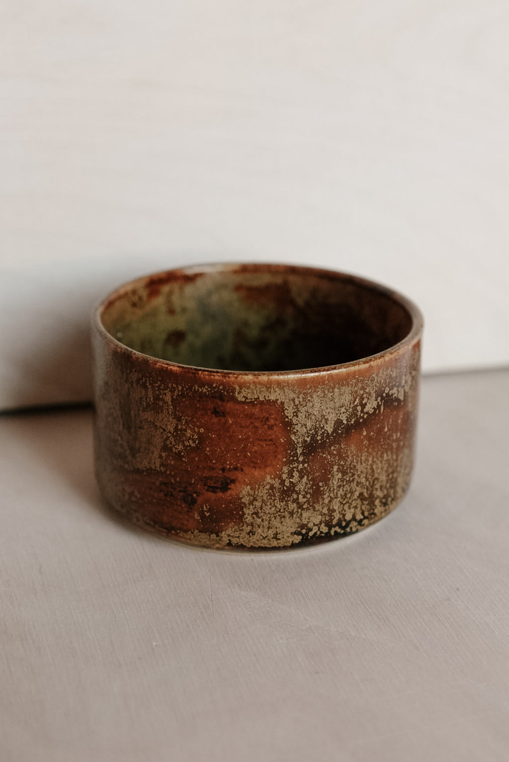 A handmade ceramic cup with a rustic, textured glaze featuring earthy brown, rust-red, and beige tones, set against a neutral light gray background.