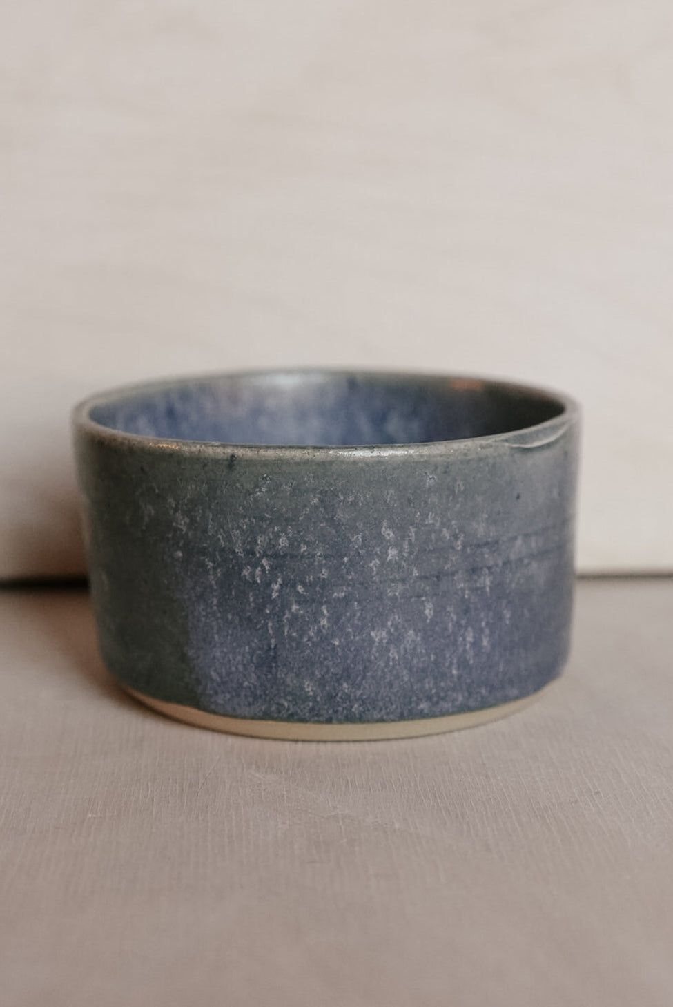 A handmade ceramic cup with a speckled slate blue and gray glaze, resting on a neutral beige surface against a light beige background.