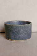 A handmade ceramic cup with a speckled slate blue and gray glaze, resting on a neutral beige surface against a light beige background.