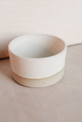 A small handmade ceramic cup with a two-tone design, featuring a matte off-white top half and a speckled beige bottom half, set against a neutral beige background.