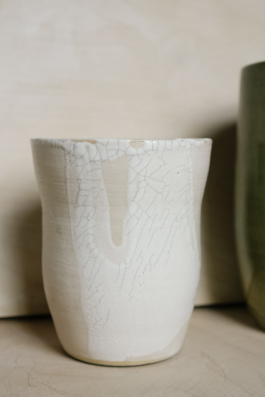A handmade ceramic vase with a textured off-white glaze featuring a crackle pattern and a vertical crack running down the center, set against a neutral beige background.