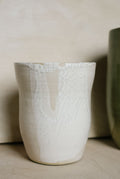 A handmade ceramic vase with a textured off-white glaze featuring a crackle pattern and a vertical crack running down the center, set against a neutral beige background.