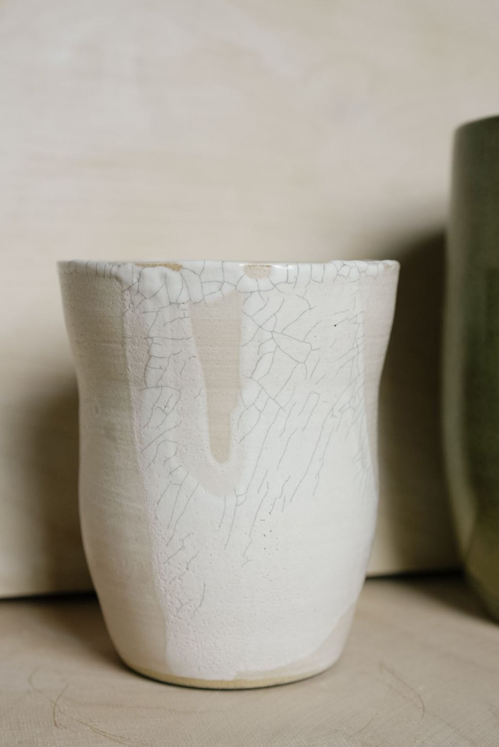 A handmade ceramic vase with a textured off-white glaze featuring a crackle pattern and a vertical crack running down the center, set against a neutral beige background.