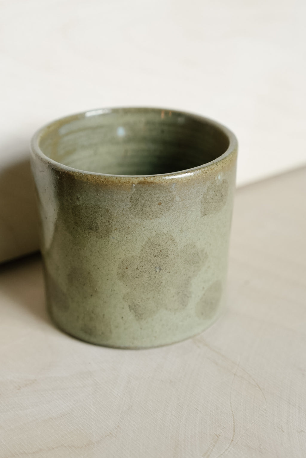 A handmade ceramic cup with a textured, speckled sage green glaze, resting on a light beige surface.