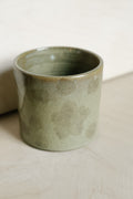 A handmade ceramic cup with a textured, speckled sage green glaze, resting on a light beige surface.