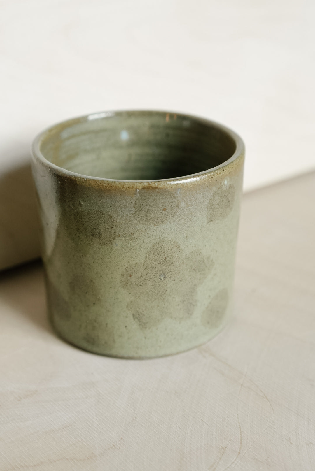 A handmade ceramic cup with a textured, speckled sage green glaze, resting on a light beige surface.