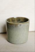 A handmade ceramic cup with a textured, speckled sage green and gray glaze, shown against a neutral off-white background.