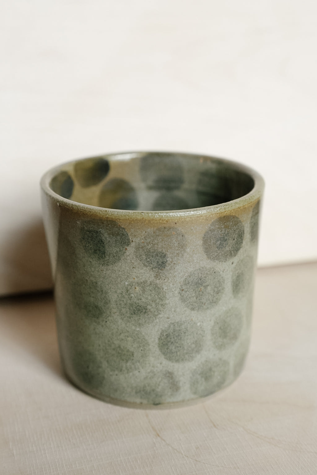 A handmade ceramic cup with a textured, hammered finish in muted sage green and gray, featuring a glossy finish and a natural unglazed rim.