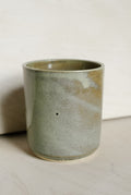 A handmade ceramic cup with a textured, speckled glaze in muted sage green and beige tones, standing on a light surface against a neutral background.