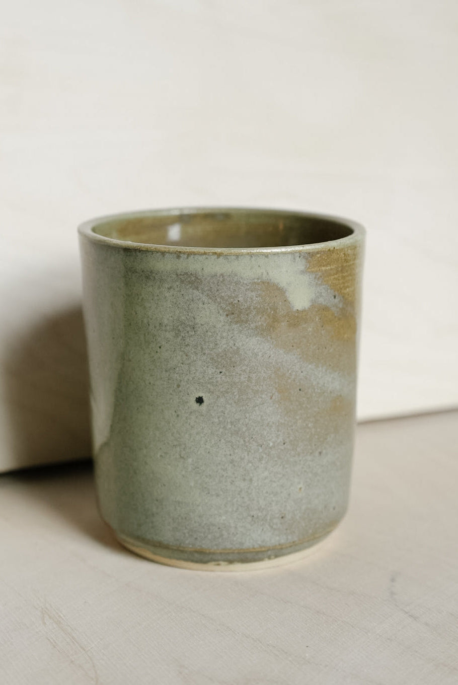 A handmade ceramic cup with a textured, speckled glaze in muted sage green and beige tones, standing on a light surface against a neutral background.