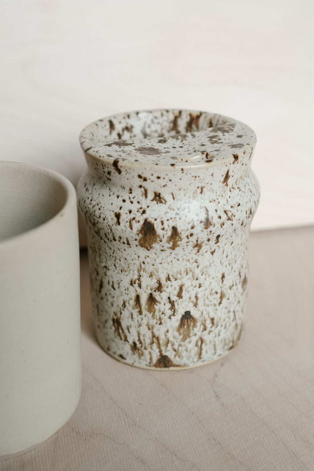 A speckled ceramic takeaway cup, featuring a textured off-white glaze with brown and beige flecks, shown next to another matte ceramic cup on a light wooden surface.