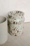 A speckled ceramic takeaway cup, featuring a textured off-white glaze with brown and beige flecks, shown next to another matte ceramic cup on a light wooden surface.