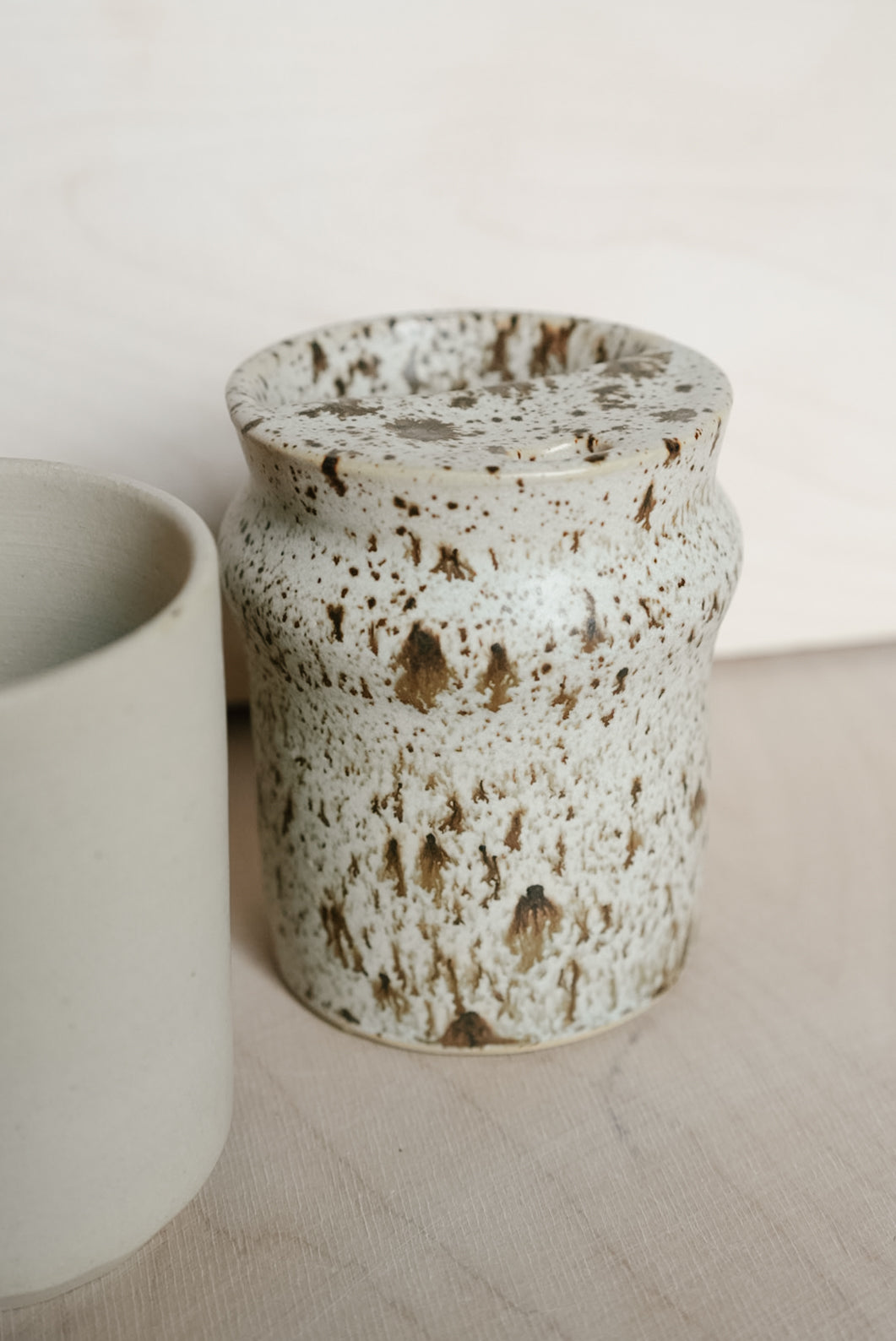A speckled ceramic takeaway cup, featuring a textured off-white glaze with brown and beige flecks, shown next to another matte ceramic cup on a light wooden surface.
