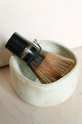 A traditional Japanese matcha whisk with a black handle and brown bamboo tines, resting inside a small, textured off-white ceramic bowl.