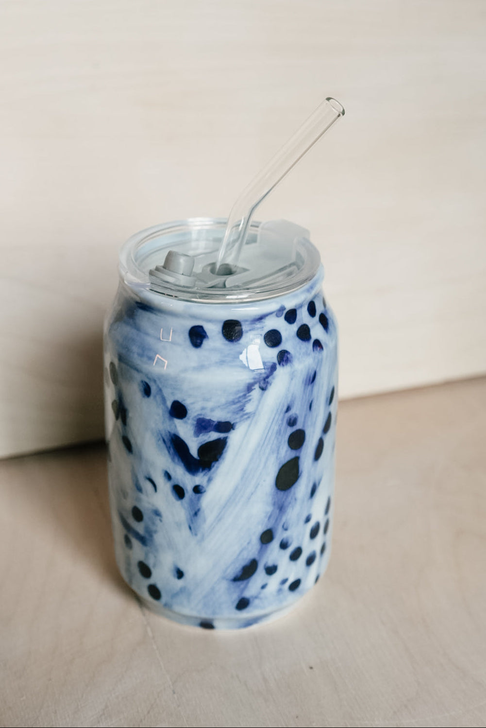 A clear glass can-shaped tumbler with a clear lid and glass straw, decorated with a blue and white marbled pattern and dark blue polka dots, set against a light wood background.