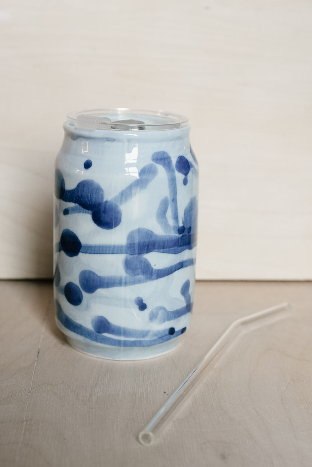 A clear plastic can-shaped straw with a clear lid, decorated with blue abstract brushstroke patterns, shown on a light wood surface against a light wood background.