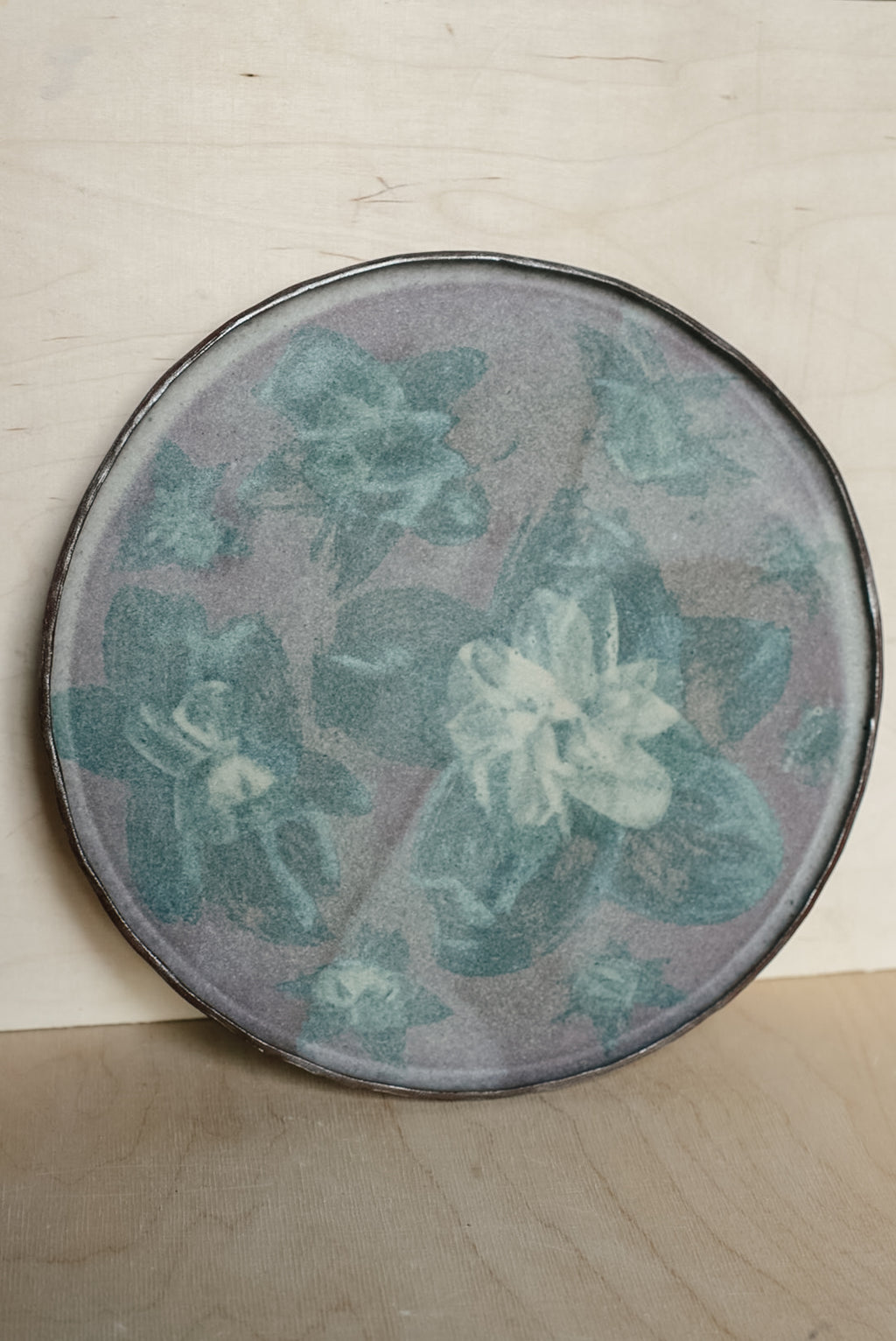 A round ceramic plate with a floral pattern in muted teal and white on a dusty purple-gray background, resting on a light wood surface.