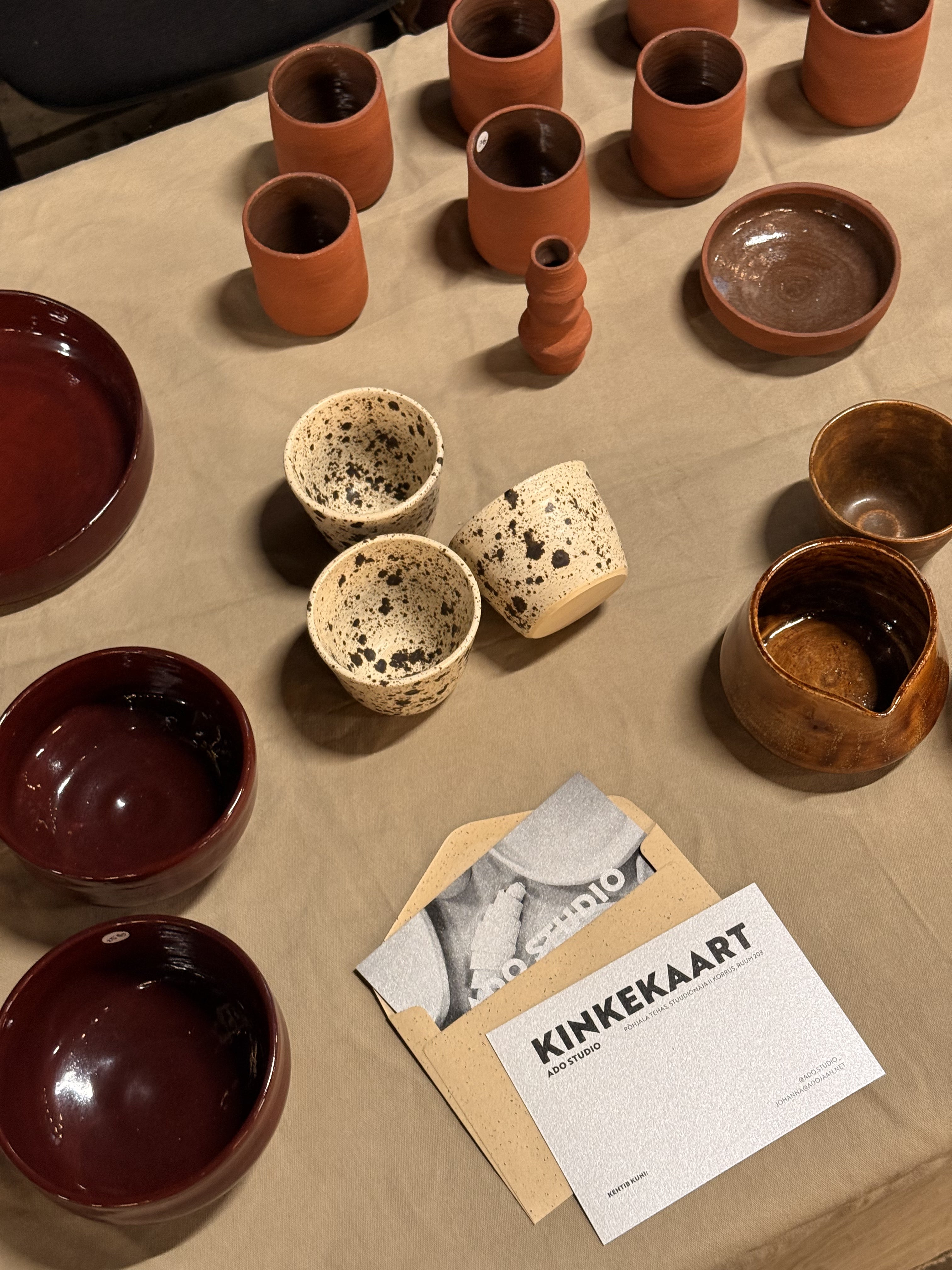 An assortment of handmade ceramic tableware, including bowls, cups, and a small vase, arranged on a beige tablecloth alongside a printed card labeled 'KINKEKAART'. The ceramics feature various glazes and finishes, including glossy deep red, speckled cream, and earthy terracotta.