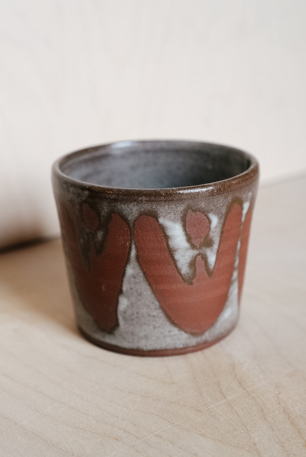 A handmade ceramic cup with a rustic design, featuring a dark brown glazed exterior with large terracotta-colored drips and a speckled gray base, set on a light wooden surface.