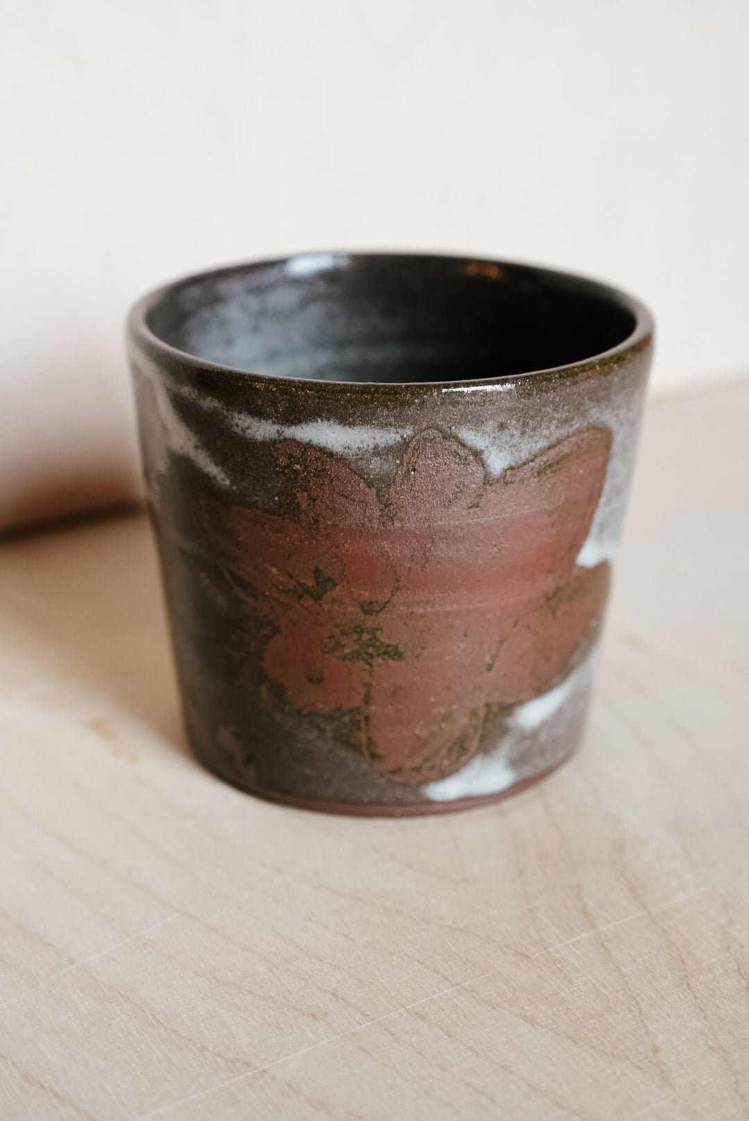 A handmade ceramic cup with a dark brown and black glaze, featuring a prominent reddish-brown floral pattern on the front and white speckled accents, resting on a light wood surface.