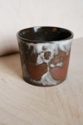 A handmade ceramic cup with a dark brown and black glaze, featuring a white and reddish-brown abstract painted pattern on the front, set on a light wood surface.