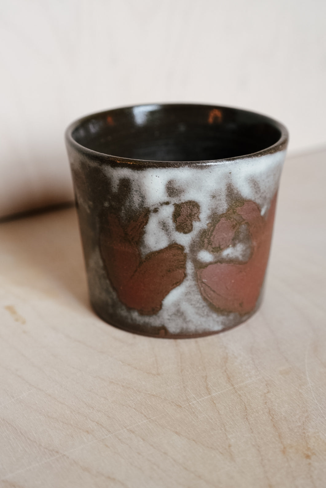 A handmade ceramic cup with a dark brown and black glaze, featuring a white and reddish-brown abstract painted pattern on the front, set on a light wood surface.