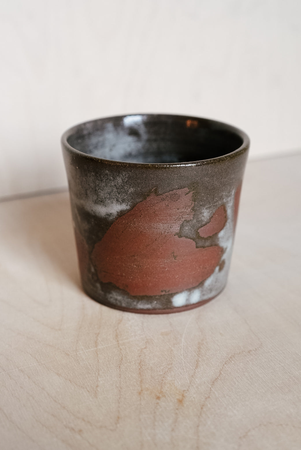 A handmade ceramic cup with a dark brown and black glaze, featuring a prominent textured reddish-brown patch on the side, set on a light wood surface against a neutral background.