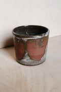 A handmade ceramic cup with a rustic finish, featuring a dark brown interior and exterior glaze with large, abstract reddish-brown and gray marbled patterns, set on a light wood surface against a neutral background.