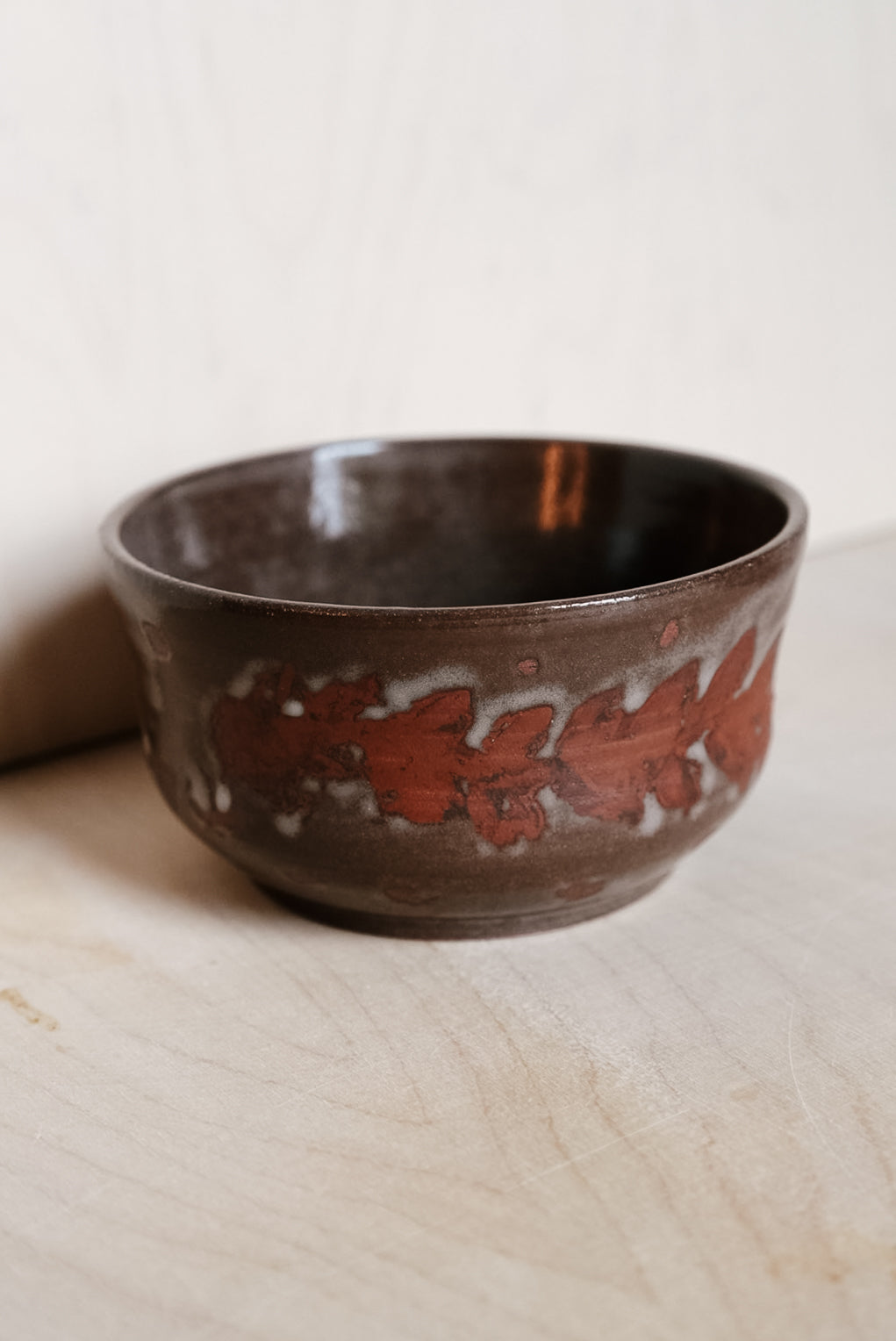 A handmade ceramic bowl with a dark brown and black glaze, decorated with a textured reddish-brown floral pattern around the middle, resting on a light wood surface against a neutral background.