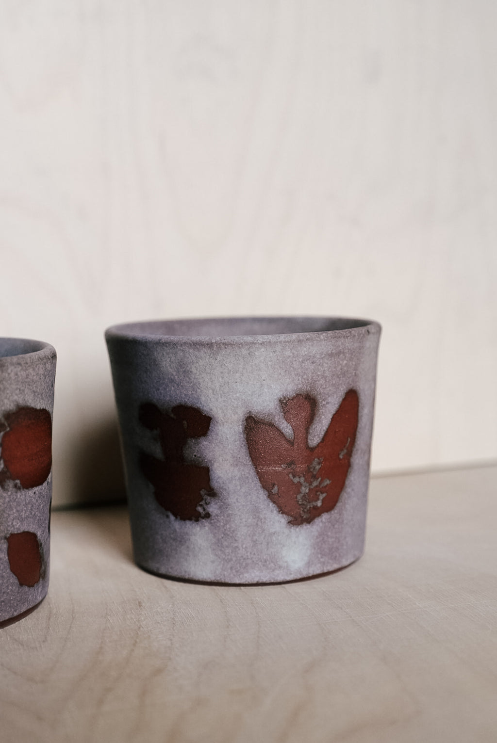A handmade ceramic cup with a textured gray and white glaze and reddish-brown abstract painted details, shown on a light wood surface against a neutral background.