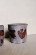 A handmade ceramic cup with a textured gray and white glaze and reddish-brown abstract painted details, shown on a light wood surface against a neutral background.